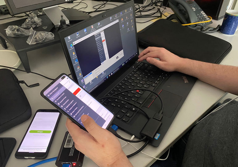 Testsituation: Laptop mit angeschlossenem Mobiltelefon, im Hintergrund das HDMI-Aufnahmegerät und einige Kabel.