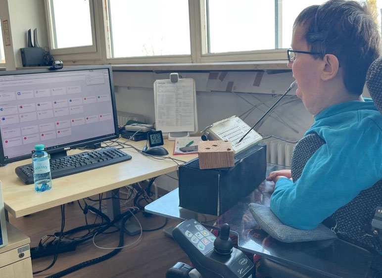 Braunhaariger Mann mit Brille, in einem Elektrorollstuhl vor einem PC sitzend, vor sich ein Podest mit einem grauen Kästchen mit Löchern.