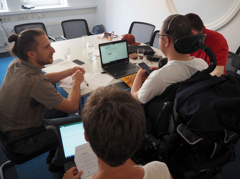 Testsituation: Testleiter, Protokollantin und Proband im Rollstuhl betrachten einen Laptop-Bildschirm